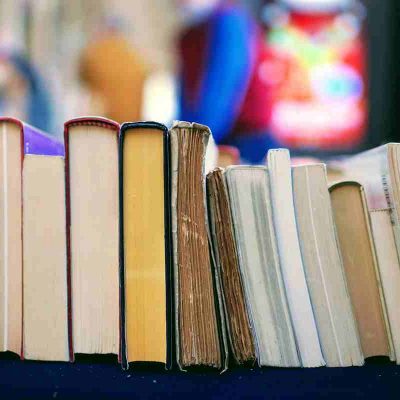 A collection of books with diverse colors and sizes stands upright on a dark surface. Behind them, the background is blurred, filled with indistinct figures and colorful shapes.