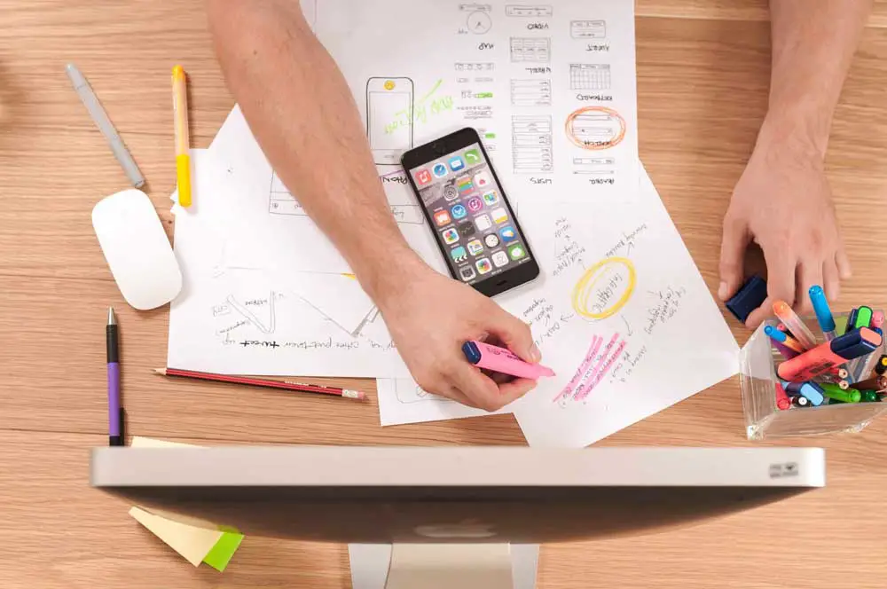 A person is diligently working at a desk filled with various design sketches, a smartphone displaying app icons, and an assortment of colored markers. With a pink highlighter in hand, they are marking up a sheet of paper. To the right side of the desk sits a container brimming with markers and other stationery items. In the foreground, there's an unmistakable presence of a computer monitor.