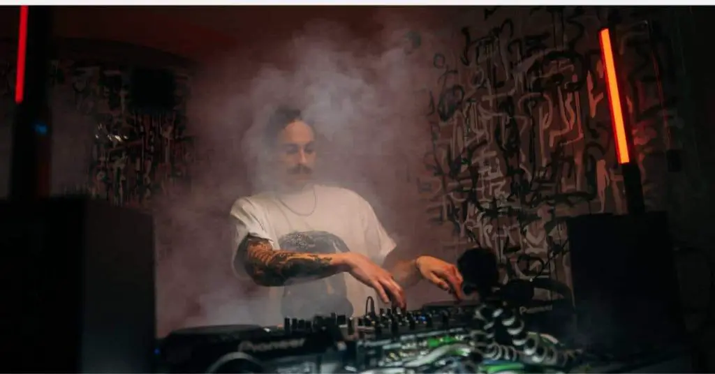 In a dimly lit room filled with fog, a DJ mixes music on turntables. Wearing a white T-shirt and showcasing tattoos on his arms, he stands against a backdrop featuring abstract graffiti art and two vertical red lights.