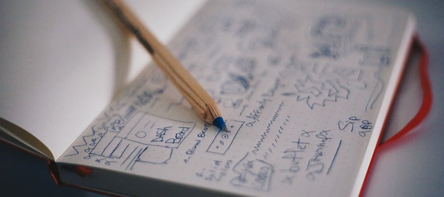 Lying on a table, an open notebook displays pages adorned with assorted sketches, doodles, and handwritten notes. Diagonally across the notebook rests a pencil, while a red ribbon bookmark peeks out from the bottom.