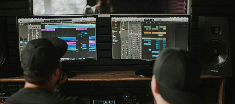 Two individuals wearing black caps are working in a music studio, seated in front of two computer monitors displaying digital audio workstation software. The setup includes speakers and various audio equipment. The room is dimly lit with a professional ambiance.