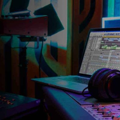 In a dimly lit music studio, a laptop displaying music production software sits on a desk. Nearby, a pair of large headphones and part of a sound mixing console can be seen. Colorful soundproofing panels and other audio equipment are visible in the background.